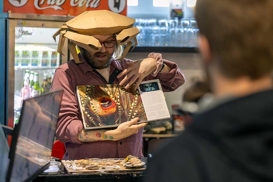 Male wearing cardboard crab hat showing a book about crustaceans at The Crab Museum in Margate, Kent