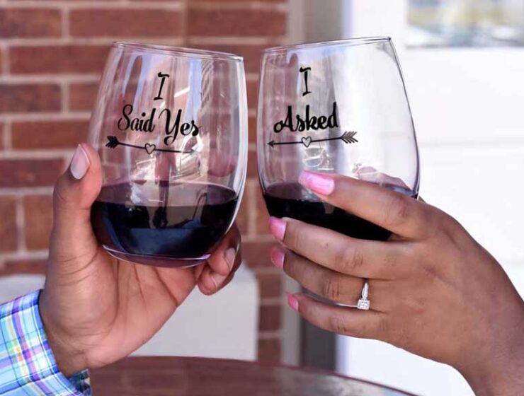 Hands holding two glasses of red wine with the words I asked, and I said yes engraved on them