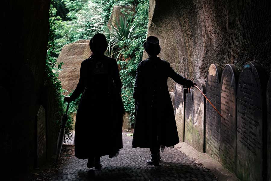 Two silhouette figures wearing top hats and carrying canes walking past tombstones in Liverpool, UK