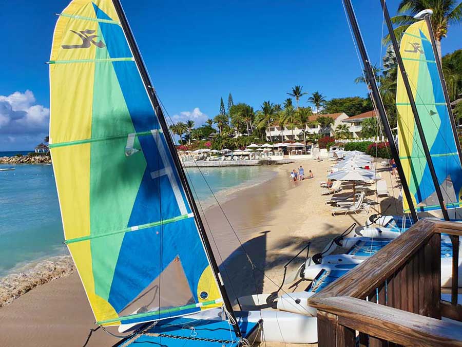 Catamarans with blue, green and yellow sails on the beach in the foreground with sun loungers and villas overlooking the bay at Blue Waters Resort Antigua