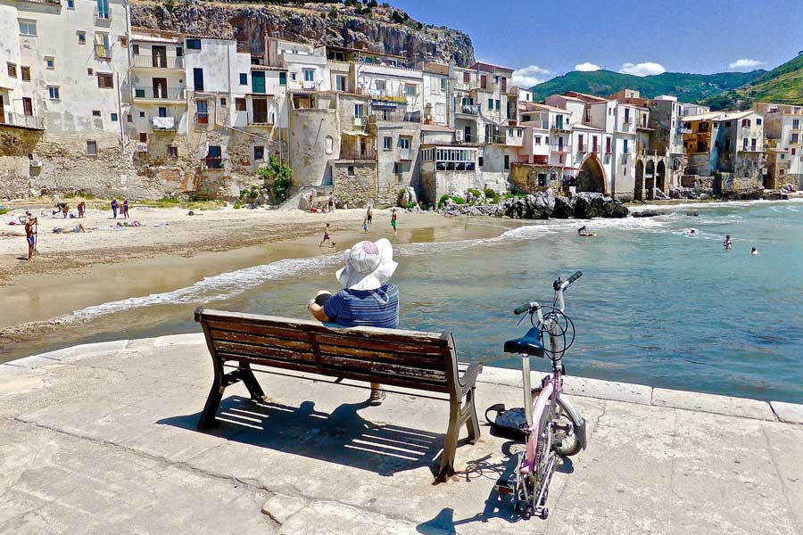 Person sitting on a bench looking across the harbour to the houses along the beach in Cefalu in Sicily.
Solo Travellers
