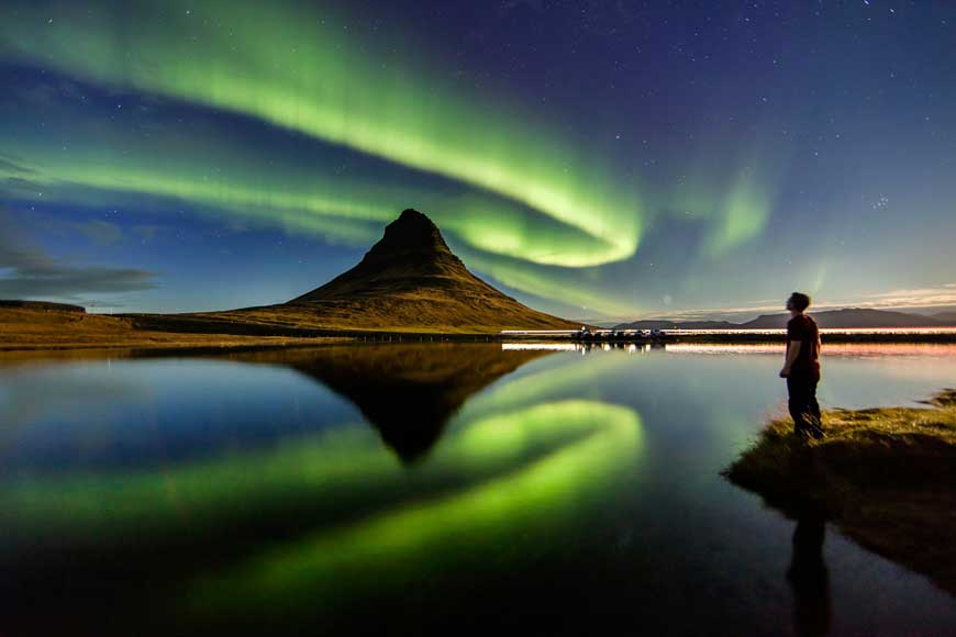 Man standing beside the body of water with Aurora lights in the sky Kirkjufell, Iceland