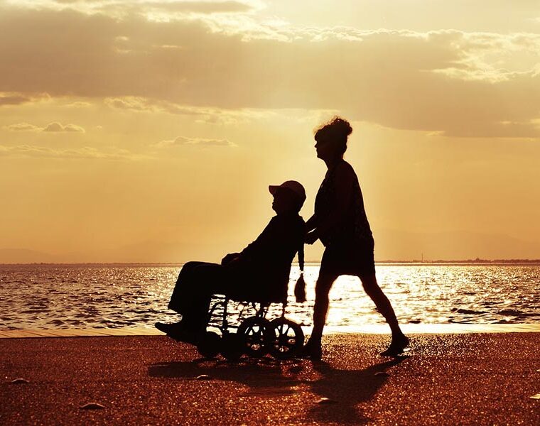 A person pushing someone in a wheelchair along the path by the sea