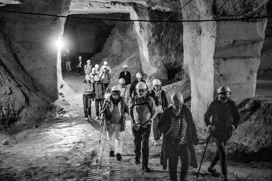 A group of visitors walks in single file through the softly lit quarry chamber, each donning yellow safety helmets and carrying backpacks or walking poles. The rough-textured walls and barrel-vaulted ceiling display the marks of hand tools used to extract stone in centuries past. Modern wiring along the ceiling brings subtle lighting to the caverns, guiding today’s guests through what was once a hive of monastic and stonemason activity.