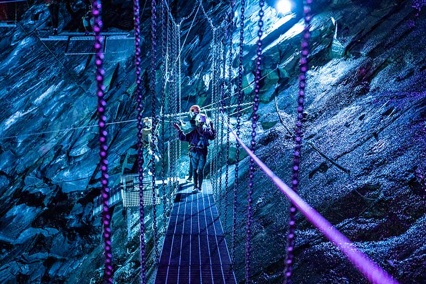 3 people wearing hard hats and safety harnesses on a walkway at the Zip World Llechwedd caverns lit by a blue light