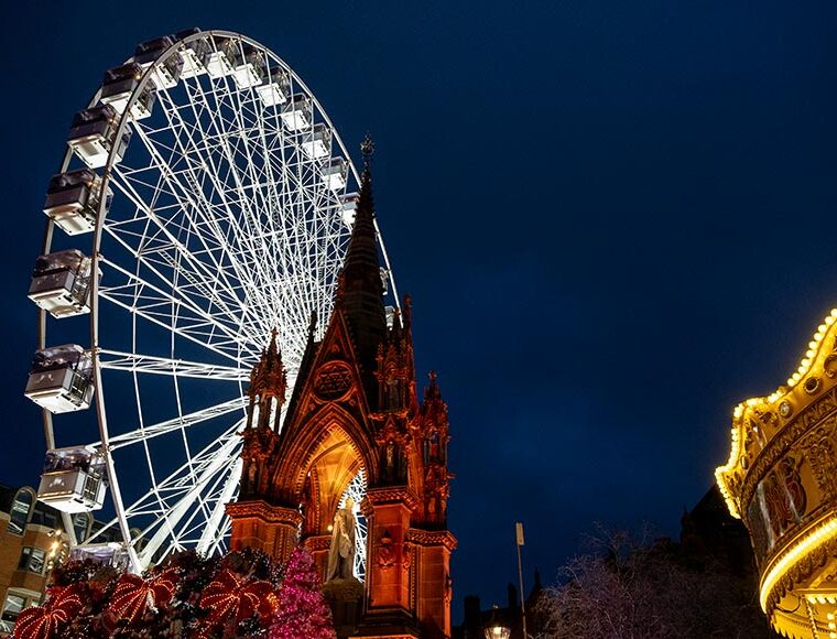 Festive setup at Albert Square in Manchester, UK, featuring the Manchester Christmas Markets with a giant Ferris wheel, an illuminated carousel, and the Prince Albert Memorial spire