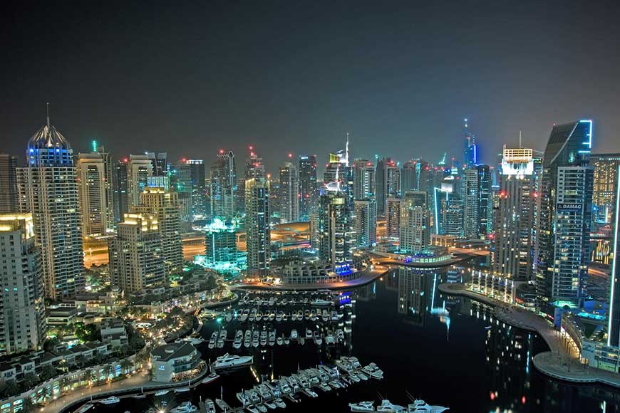 The illuminated skyline of Dubai Marina at night, featuring numerous skyscrapers and a marina filled with yachts.