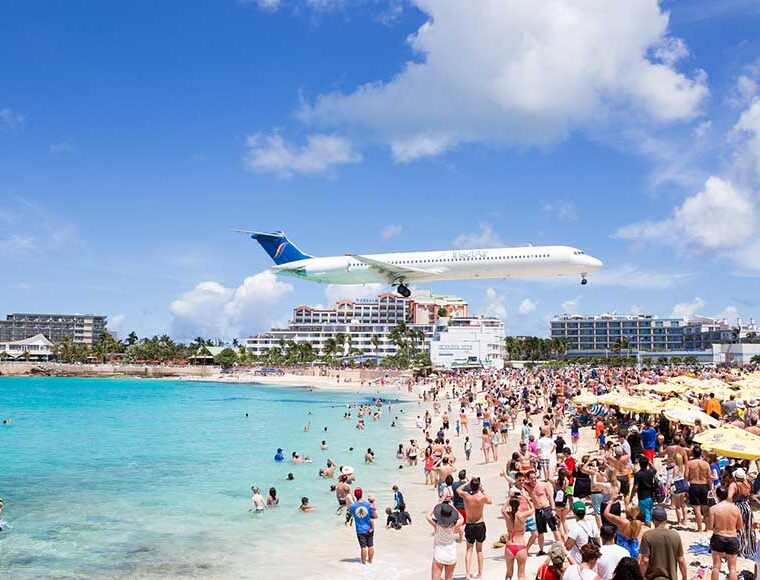Aircraft flying across a crowded beach at Maho Beach in Sint Maarten approaching the runway at Princess Juliana International Airport.