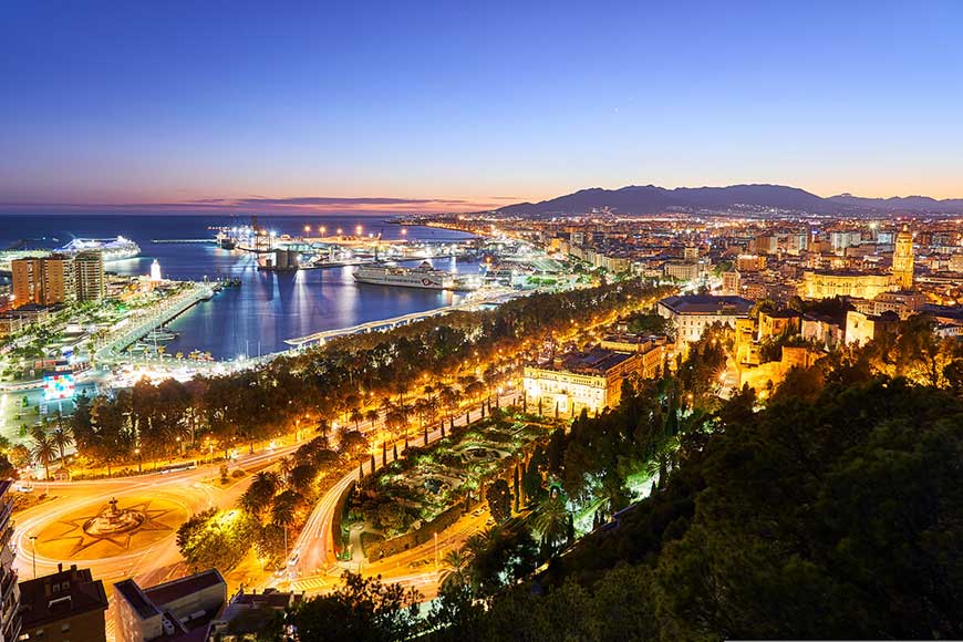 A panoramic view of Málaga, Spain, at dusk, featuring the illuminated city and its harbour, with the prominent Castillo de Gibralfaro in the foreground