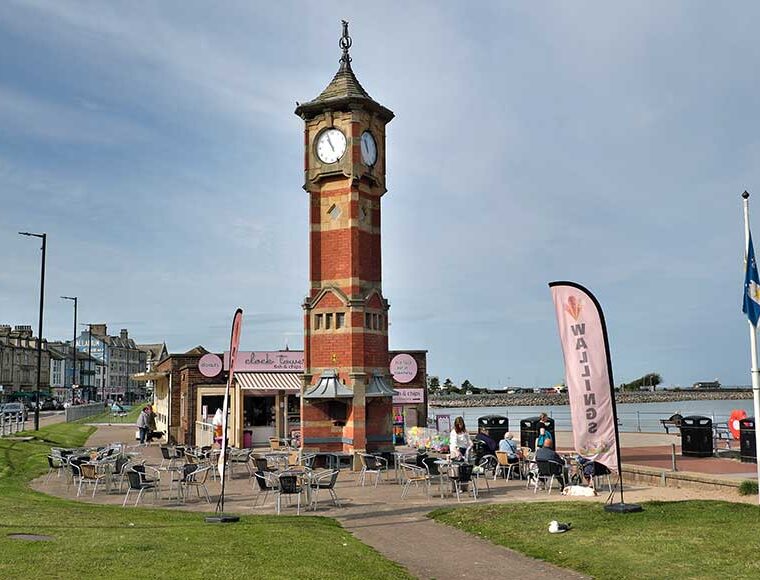 The Clock Tower Morecambe made of red brick with sandstone banding and dressings. Of square plan above a cruciform base with corner buttresses. Behind is the Clock Tower Café and Morecambe Promenade