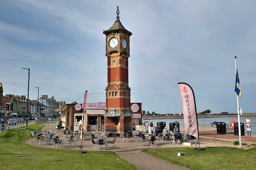The Clock Tower Morecambe made of red brick with sandstone banding and dressings. Of square plan above a cruciform base with corner buttresses. Behind is the Clock Tower Café and Morecambe Promenade