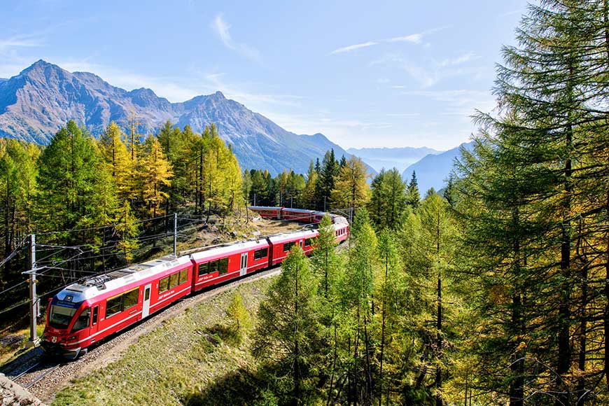 Red passenger train curves through a forested mountain landscape on a sunny day.