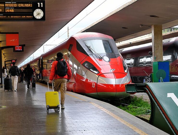 Red high-speed train at a platform with passengers boarding; a man with a yellow suitcase in the foreground.