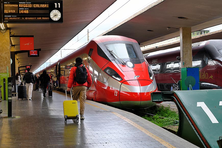 Red high-speed train at a platform with passengers boarding; a man with a yellow suitcase in the foreground.