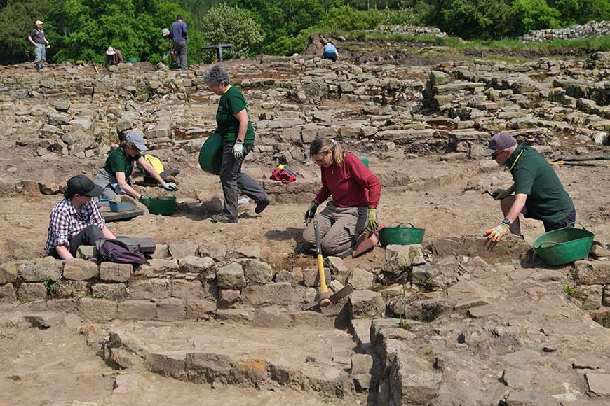 Archaeologists excavating a stone ruin site, kneeling and digging with trowels and buckets.