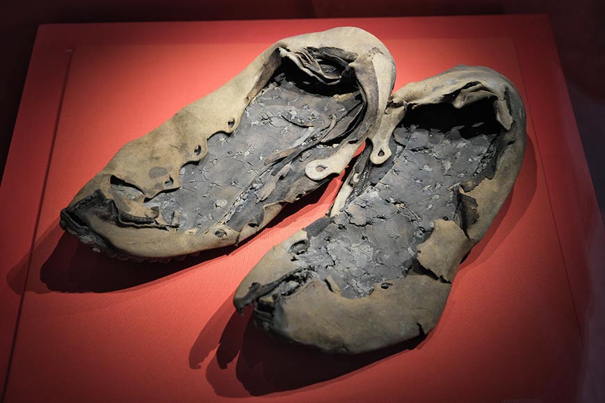 Pair of ancient, charcoal-blackened leather shoes on a red display background in a museum case, preserved as artifacts.