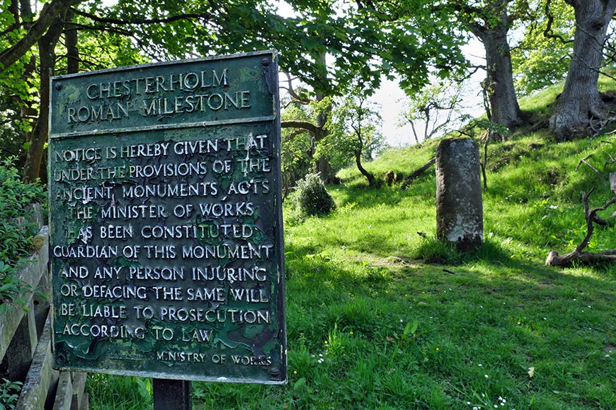 Weathered sign reading Chesterholm Roman Milestone amid a grassy, shaded hillside with trees behind it.