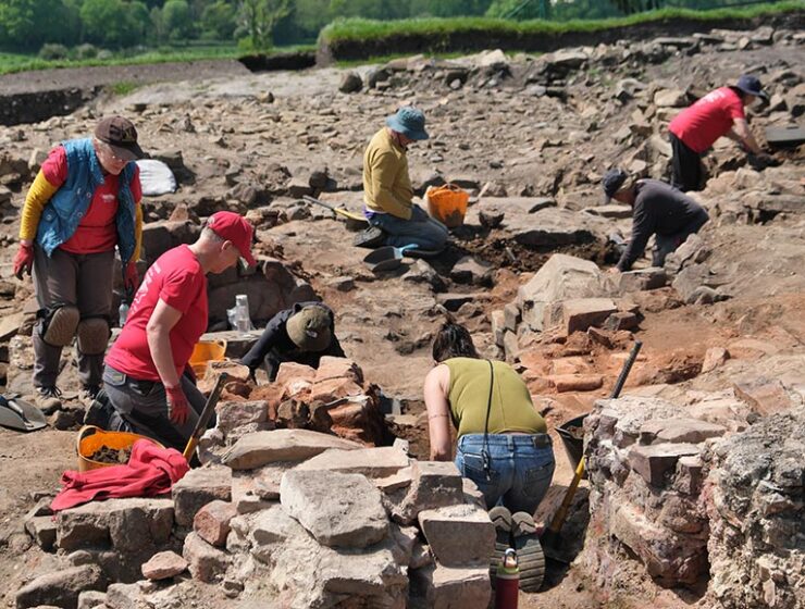People excavating at an outdoor archaeological dig site, sorting and moving rocks with trowels and buckets.