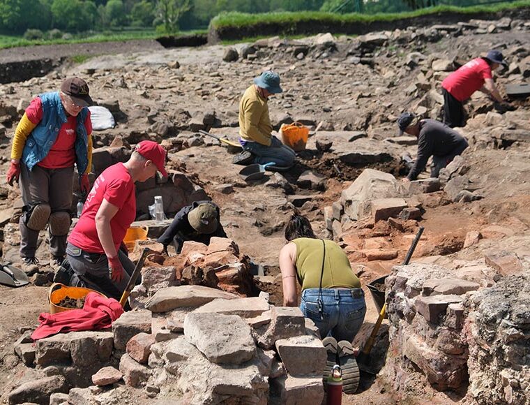 People excavating at an outdoor archaeological dig site, sorting and moving rocks with trowels and buckets.