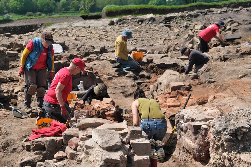 People excavating at an outdoor archaeological dig site, sorting and moving rocks with trowels and buckets.