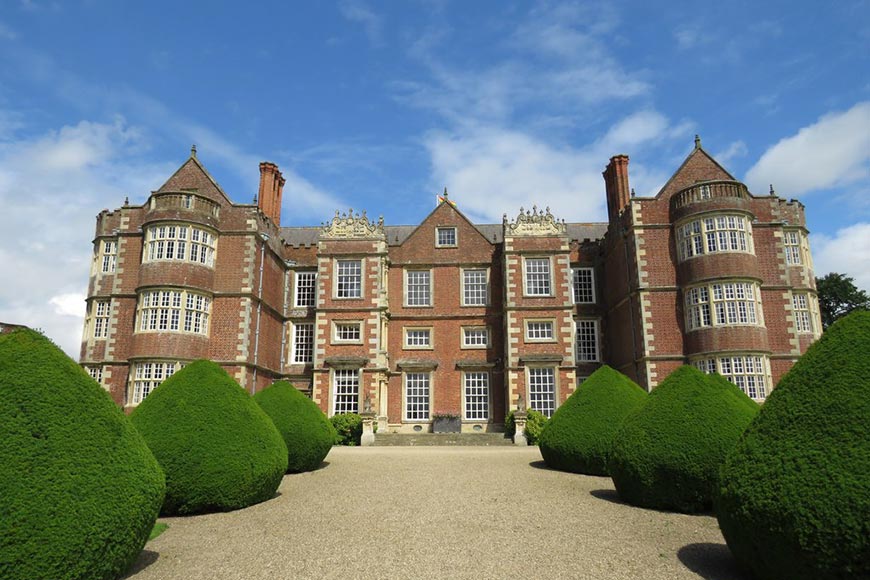 Stately red-brick manor with tower-like wings, decorative stonework, and a gravel path framed by neatly clipped cone-shaped hedges.