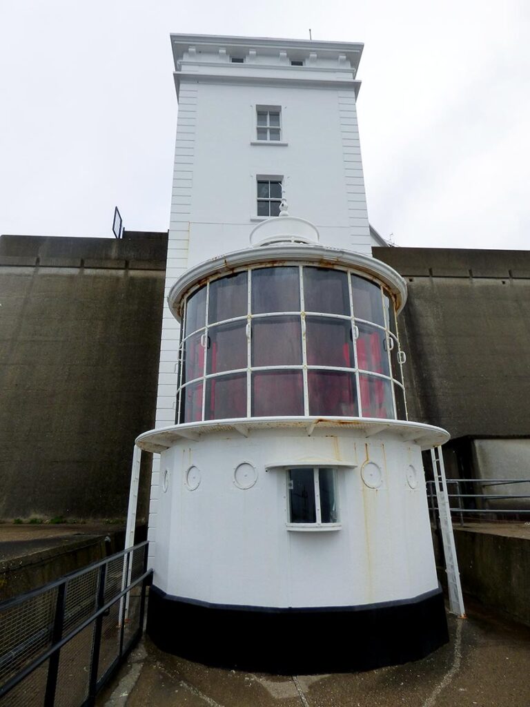 White lighthouse-like tower with a cylindrical glass observation room at the base and rust stains, set against a gray concrete wall.