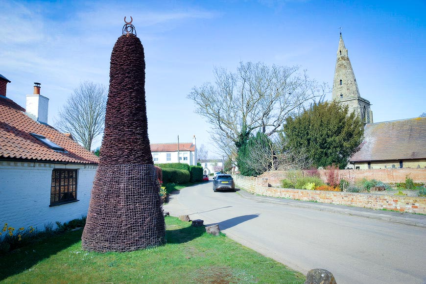 Cone-shaped wicker sculpture on a grassy verge beside a white cottage in a quiet village street with a blue sky above.