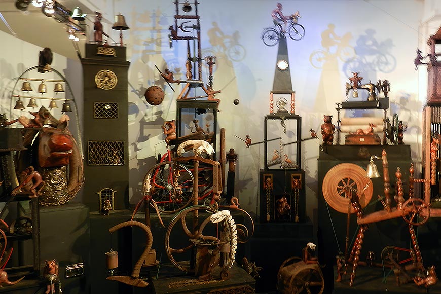 Eclectic museum display of antique mechanical toys and devices—bicycles, clocks, gears, and wooden figures on shelves and pedestals with shadows on a blue wall.