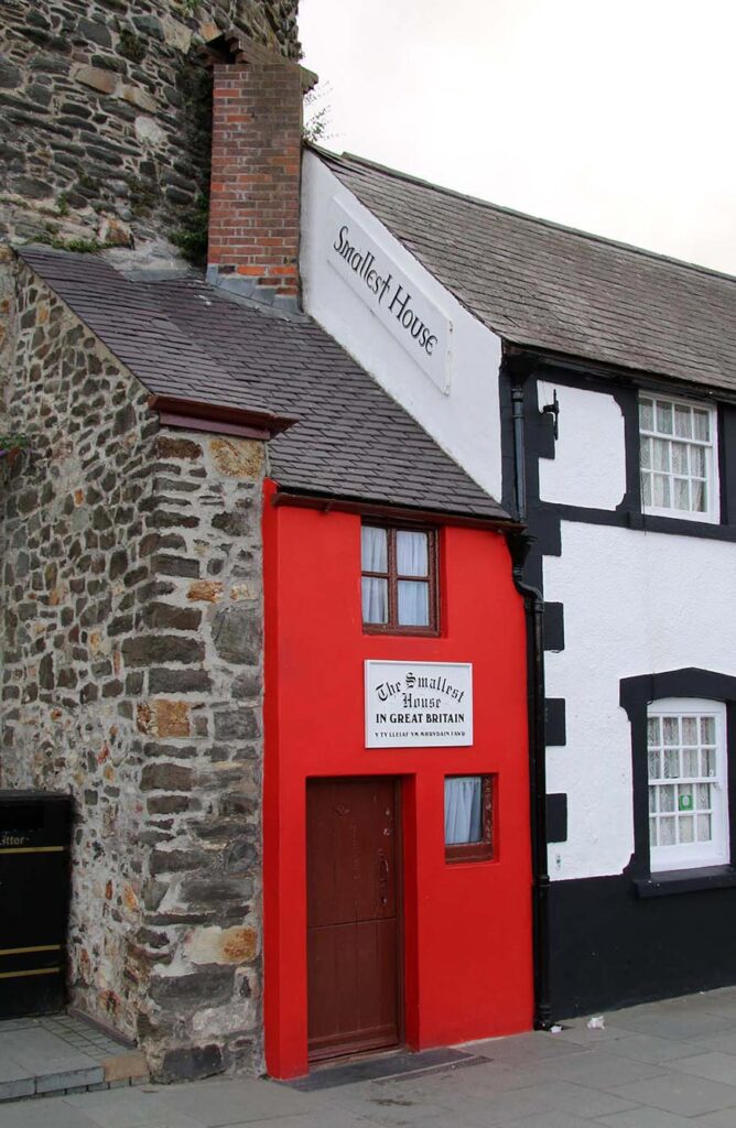 Red painted house labelled 'The Smallest House in Great Britain' between a stone wall and a black-and-white building.