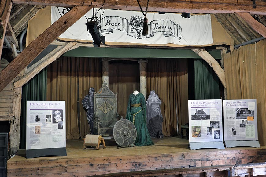 Stage in a rustic barn theatre with period costumes on mannequins and two large informational panels on either side of the stage.