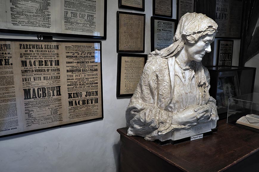 Marble bust of Ellen Terry in period clothing on a wooden pedestal, beside vintage theatre posters on the wall with plaques nearby.