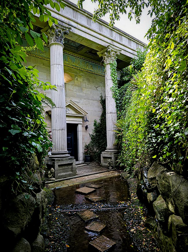 The image shows a classical temple facade set within a sunken water feature surrounded by rock walls and ferns at The Forbidden Corner in North Yorkshire, England.