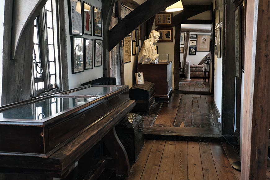Wooden hallway of a historic house with a bust of Ellen Terry, framed photos on the walls, and a glass display case to the left.