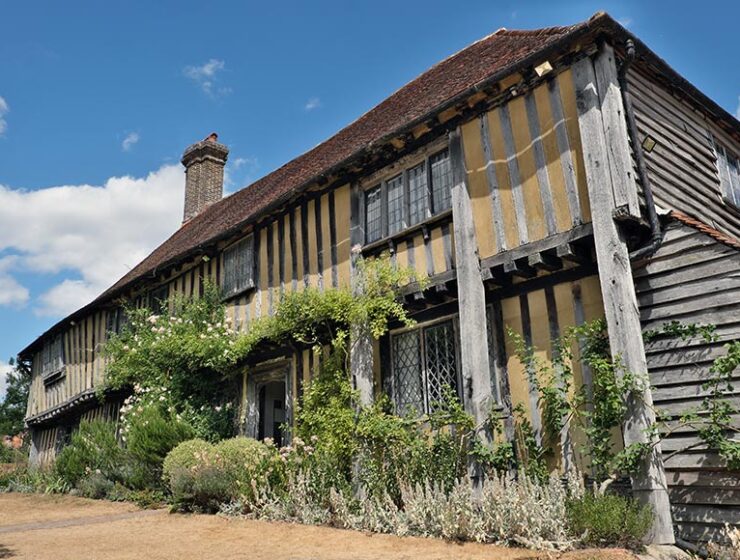 Old half-timbered cottage with weathered wood and a lush, overgrown garden under a blue sky.