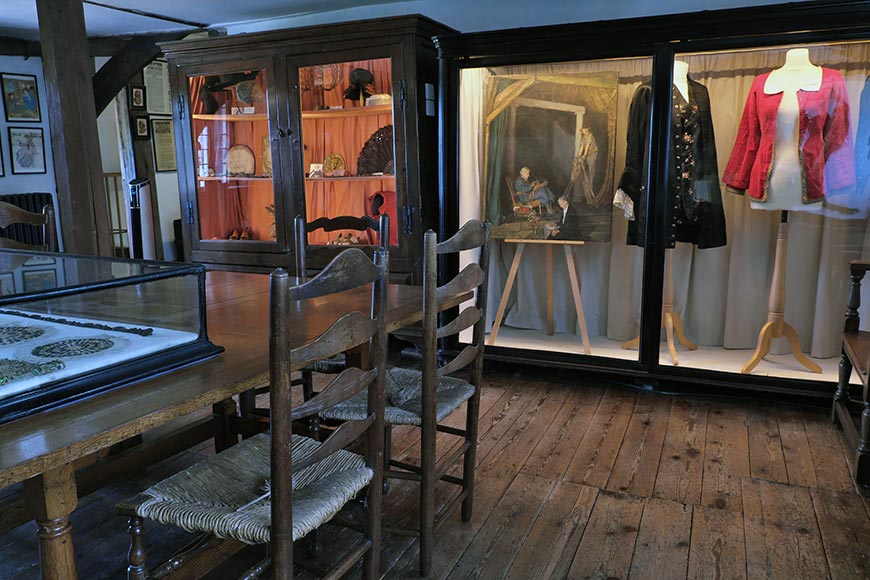 Ellen Terry museum exhibit with wooden chairs around a large display table and glass cases showing clothing and artifacts on shelves and mannequins in the background