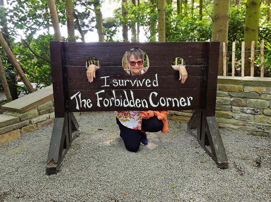 Visitor posing with her head and hands locked in a wooden pillory reading "I survived The Forbidden Corner" at the famous folly garden in Tupgill Park, North Yorkshire